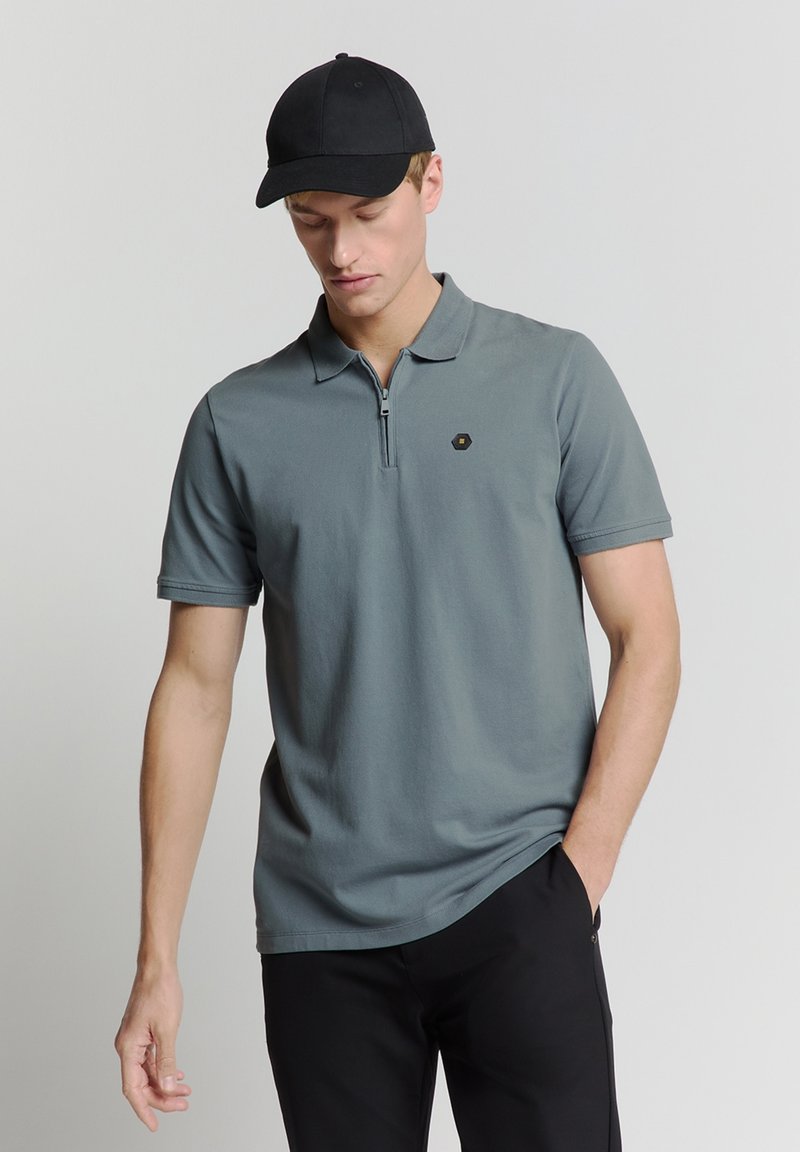 Grijze polo met korte mouwen, een zip-kraag en een klein zwart logo, gecombineerd met een zwarte pet en broek. Gladde textuur.