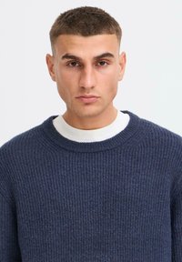 Jeune homme avec des cheveux bruns courts portant un pull en tricot bleu marine sur une chemise blanche, regardant vers l'avant avec une expression neutre.