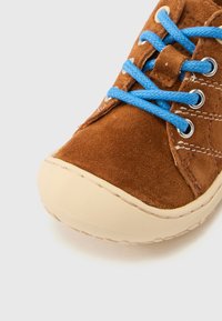 Καφέ παπούτσι από σουέντ με μπλε κορδόνια, στρογγυλεμένη μύτη και μπεζ καουτσούκ σόλα. Διαθέτει ραμμένες λεπτομέρειες και σύστημα κορδονιών με κορδονότρυπες.