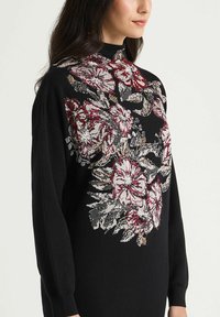 Pull noir avec motif floral en rouge, rose et crème. Col montant, manches longues et tissu tricoté texturé.