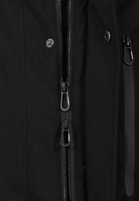 Schwarze Jacke mit mehreren Reißverschlüssen, einem Druckknopf und einer seitlichen Reißverschlusstasche, aus einem glatten, strapazierfähigen Stoff.
