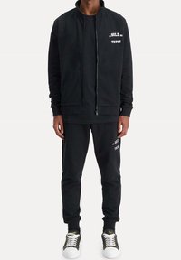 Černá mikina a tepláky vyrobené z měkké tkaniny. Bundě na zip dominuje bílý text; kalhoty mají odpovídající textový detail. Doplněno teniskami.