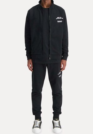 Black sweatshirt and sweatpants made of soft fabric. Zippered jacket features white text; pants have matching text detail. Accented with sneakers.