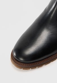 Botín de cuero negro hasta el tobillo con punta redondeada y acabado liso. Presenta una suela cosida en marrón, destacando un diseño clásico.