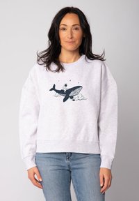 Vrouw met donker haar die een lichte sweatshirt draagt met een grafische afbeelding van een astronaut die op een walvis rijdt, omringd door wolken en sterren.