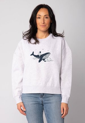 Frau mit dunklen Haaren, die einen hellen Sweatshirt trägt, das ein Grafikdesign eines Astronauten zeigt, der auf einem Walfisch reitet, umgeben von Wolken und Sternen.