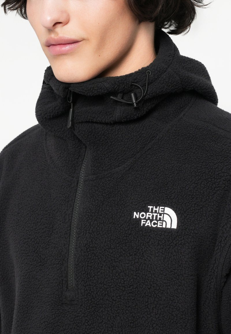 Eine Person trägt eine schwarze Fleecejacke mit hochschließendem Reißverschluss und dem weißen The North Face-Logo auf der Brust vor einem einfarbigen Hintergrund.