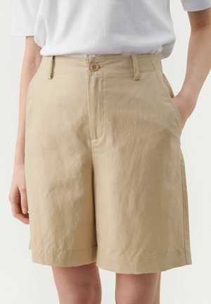 Pantalones cortos de talle alto beige con cierre de botón, trabillas para cinturón, bolsillos laterales, usados con una camiseta blanca de manga corta metida por dentro.