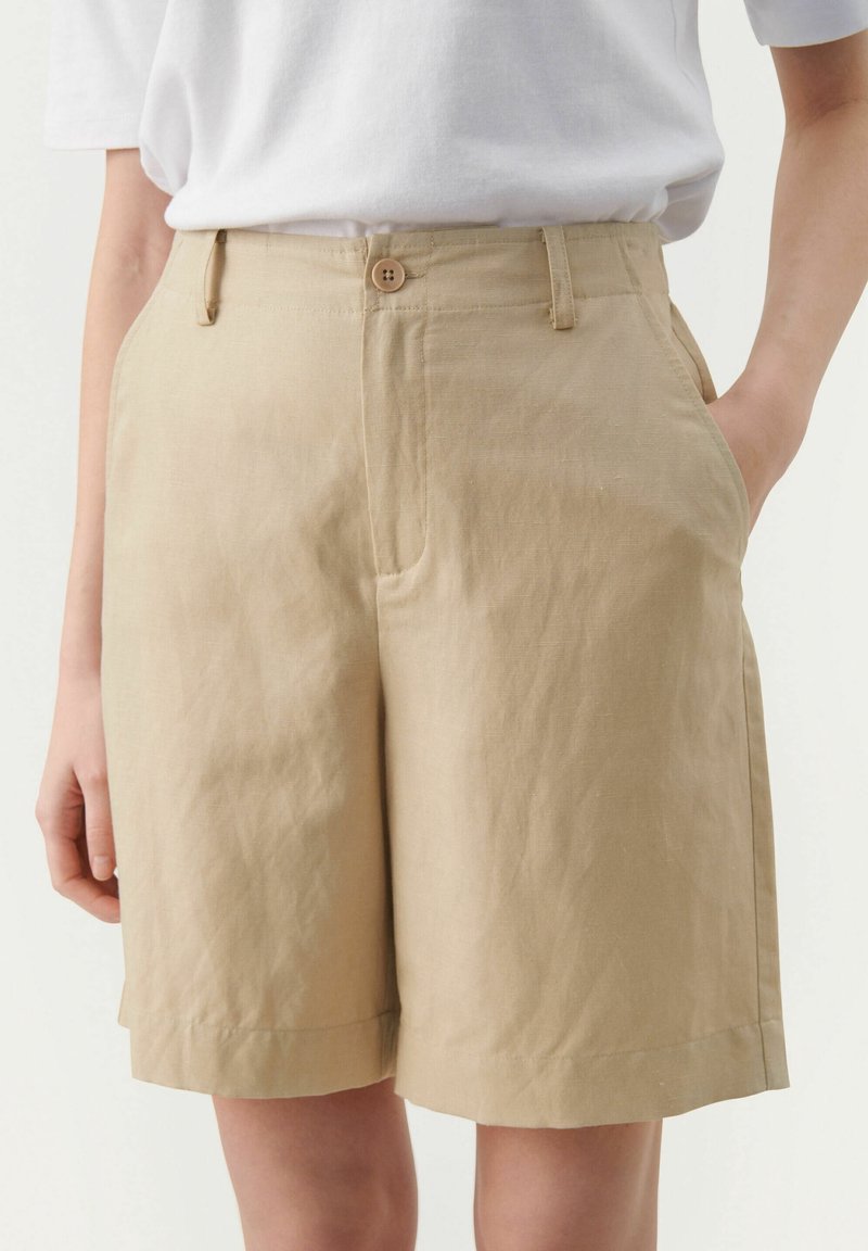 Short beige taille haute avec fermeture par boutons, passants pour ceinture, poches latérales, porté avec une chemise blanche à manches courtes rentrée.
