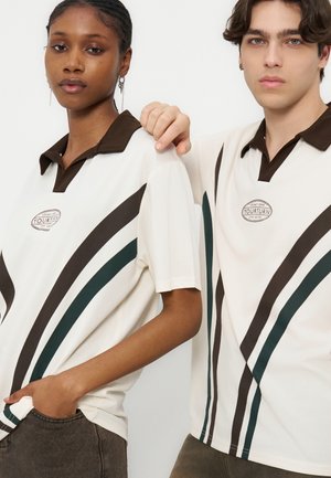 Két fiatal felnőtt, akik ugyanolyan fehér, sötét galléros, átlós barna és zöld csíkos pólót viselnek, az egyik a másik vállára teszi a kezét.