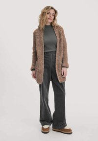 Cardigan en tricot texturé et aéré beige, porté par-dessus un haut vert foncé, associé à un pantalon en velours côtelé gris à jambes larges et des bottes marron doublées en fourrure.