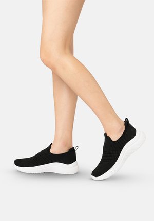 Nogi w czarnych dzianinowych slip-onach z białymi grubymi podeszwami, jedna stopa stawiająca krok do przodu na jasnoszarym tle.