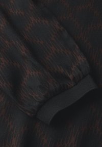 Gros plan sur une manche en tissu foncé avec un motif en zigzag noir et marron subtil, et un poignet élastique noir.