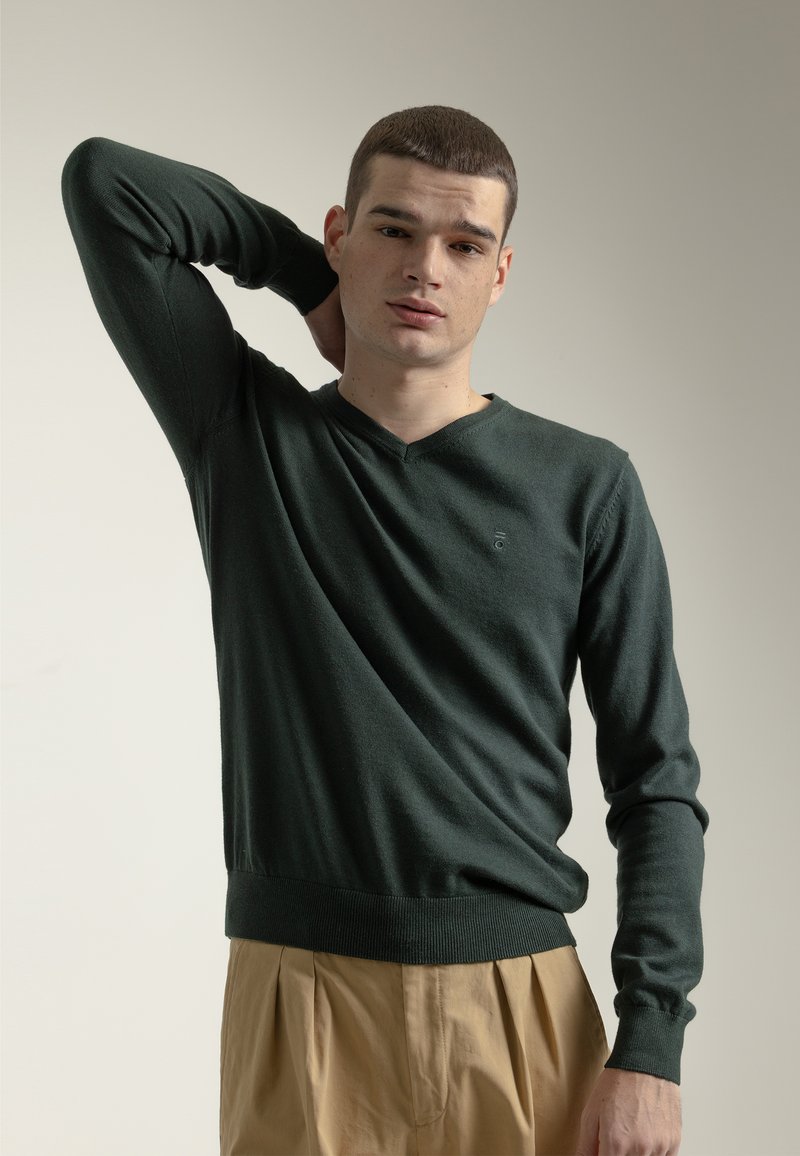 Suéter de punto verde oscuro con escote en V, puños y dobladillo de canalé. Textura suave con un pequeño logo en el pecho. Combinado con pantalones de color claro.