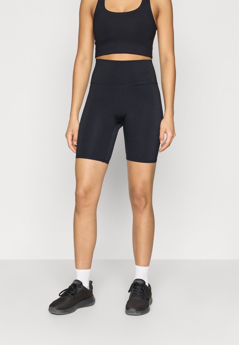 Top atletico nero crop e pantaloncini da bici a vita alta. Tessuto liscio ed elastico con design aderente e lunghezza a metà coscia, abbinati a sneakers nere.