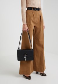 Borsa a tracolla in pelle nera con catena argentata e chiusura a patta, caratterizzata da un accentuazione del logo metallico e forma rettangolare.