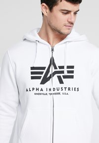 Biela mikina na zips s voľným strihom, s čiernym grafickým logom na hrudi. Tkanina vyzerá mäkko a má šnúrky pri kapucni.