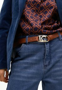 Brązowy skórzany pasek z srebrną klamrą z logo, noszony na ciemnoniebieskich dżinsach. Wzorzysta koszula z pomarańczowymi i granatowymi powtarzającymi się literami. Granatowa marynarka.