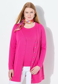 Cárdigan fucsia de manga larga con botones plateados, combinado con una blusa a juego. Presenta una bufanda de patrón colorido en el cuello.