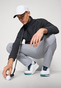 Un hombre con un suéter negro con cremallera y pantalones grises se agacha, sujetando una pelota de golf cerca de un palo de golf negro. Lleva zapatos de golf blancos y azules y una gorra blanca.