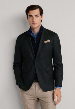 Blazer noir sur un pull marine avec un col zippé, associé à une chemise bleu clair, complété par un pantalon beige et un carré de poche à motifs.