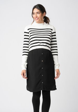 Smilende kvinde med mørkt hår i en hestehale iført en hvid og sort stribet sweater, sort knappet nederdel og sorte leggings, der står på en hvid baggrund.
