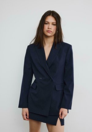 Jonge vrouw met steil donker haar, gekleed in een marineblauwe dubbelrijige blazer met krijtstreep en een bijpassende rok met een voorwaartse split.