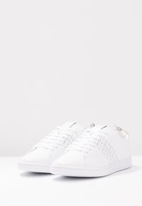 Zapatillas blancas con un upper texturizado que presenta el logotipo repetido de "Lacoste", puntera redondeada, suela plana y un acento plateado en el talón.