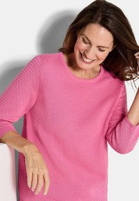 Eine lächelnde Frau, die nach unten schaut, trägt einen strukturierten pinken Pullover, mit einer Hand, die ihr Haar berührt, und der anderen Hand, die auf einer Oberfläche ruht.