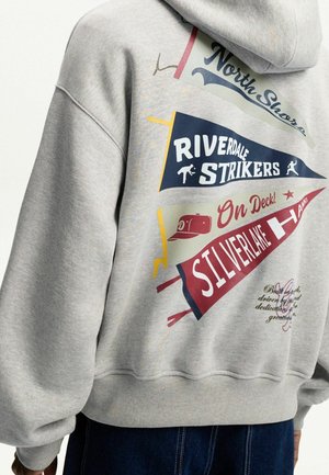 Šedá mikina s kapucí s barevnými praporky nesoucí nápisy "North Shore", "Riverdale Strikers", "On Deck" a "Silverlake Hawks" a s malými ikonami tématicky spojenými s baseball.