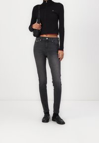 Černý žebrovaný zipový top s červenými akcenty je doplněn o světle šedé skinny džíny s vysokým pasem. Modelka nosí černé mokasíny. Jednoduchý, přiléhavý design.