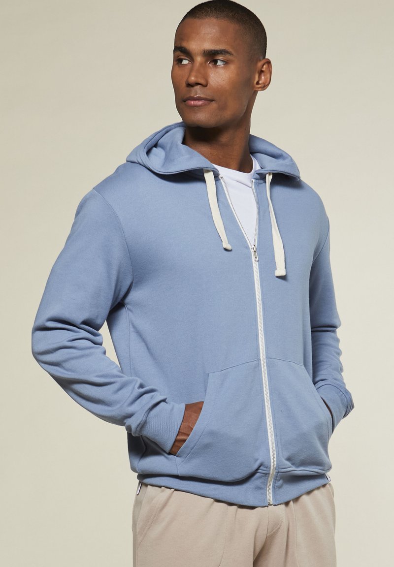 Hellblaue Zip-Hoodie mit einer Fronttasche, weißen Kordeln und gerippten Bündchen. Hergestellt aus weichem Material mit lockerem Schnitt.