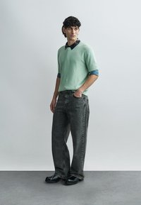 Miętowy, teksturowany sweter z krótkim rękawem, nałożony na czarną koszulę z kołnierzykiem, w połączeniu z luźnymi szarymi dżinsami i czarnymi butami.