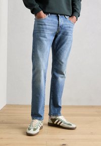 Lichtblauwe denim spijkerbroek met een rechte pasvorm, voorzien van vijf zakken en een vervaagde afwerking. Gecombineerd met groene en grijze sportschoenen.