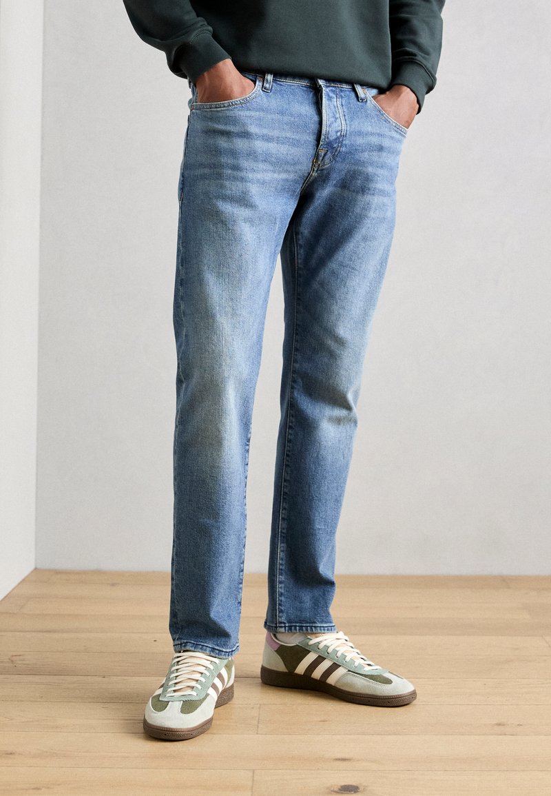 Lichtblauwe denim spijkerbroek met een rechte pasvorm, voorzien van vijf zakken en een vervaagde afwerking. Gecombineerd met groene en grijze sportschoenen.