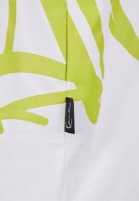 White cotton t-shirt featuring bold lime green abstract patterns. Includes a small black label with a white graphic detail.