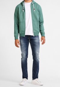 Sudadera verde con cremallera y cordones, con bolsillos frontales, llevada sobre una camiseta gris. Jeans azul con ligero desgaste y zapatillas blancas.