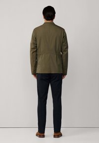 Giacca leggera verde oliva con una vestibilità rilassata, caratterizzata da una ventola posteriore e due tasche laterali inferiori, abbinata a pantaloni scuri a sigaretta e scarpe marroni.