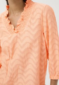 Ferskenfarvet top med flæsekant ved halsudskæringen og 3/4 ærmer. Har et struktureret zigzag-mønster. Fremstillet af letvægtsstof.