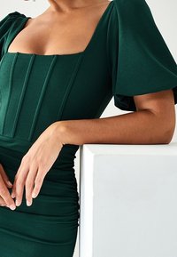 Robe ajustée vert foncé avec un décolleté carré, un corsage plissé et des manches bouffantes ; tissu texturé, détails froncés à la taille.