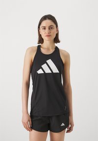 Černý sportovní top bez rukávů vyrobený z hladkého materiálu, s velkým bílým logem Adidas uprostřed. Design má tenké ramínka a kulatý výstřih.