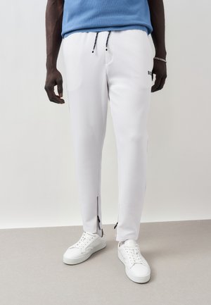 Férfi fehér slim fit jogger nadrág oldalán cipzárakkal a bokánál, fehér tornacipőkkel és kék pulóverrel párosítva.