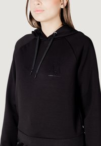 Czarna bluza z kapturem o skróconym kroju, teksturowanej tkaninie, tonalnym logo i sznurków ściągających. Miękka i dopasowana w pasie.