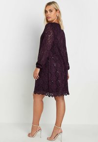 Vestido de encaje en un profundo tono púrpura con mangas largas, dobladillo festoneado y acentos de lentejuelas. Combinado con tacones plateados con tiras, mostrando una textura delicada.