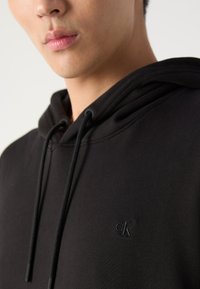 Čierna mikina z mäkkého materiálu, s kapucňou na šnúrku a malým embosovaným logom "CK" na hrudi.