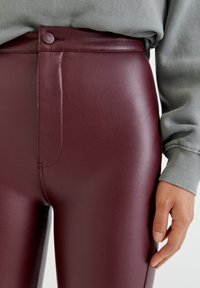 Nära bild av en person som bär högmidjade, vinröda byxor i faux läder och en grön ribbad långärmad topp med avslappnade manschetter.