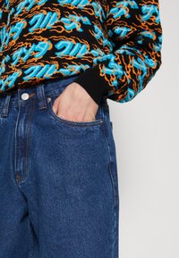 Sweater de malha multicolorido com padrões preto, azul e laranja, combinado com jeans de denim azul com fecho de botão e bolsos laterais.