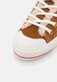 Brązowe zamszowe trampki z białymi sznurówkami i gumowym noskiem. Posiadają puszystą teksturę w okolicy sznurówek oraz czerwony akcent na podeszwie.