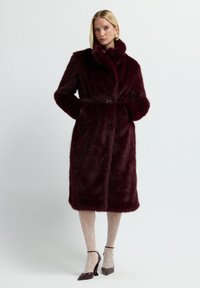 Nainen, jolla on ylleen puettu polvipituinen tumman punainen tekoturkiskääntö, vyö, valkoiset pitsi sukkahousut ja punaiset korkokenkä, seisoo valkoista taustaa vasten.