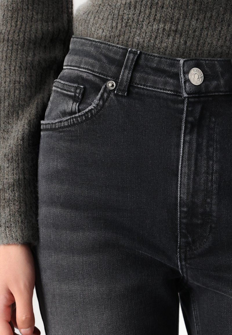Jeans neri in denim a vita alta con una texture liscia, design a cinque tasche e dettaglio in metallo argento vicino al girovita, abbinati a un maglione grigio a maglia.